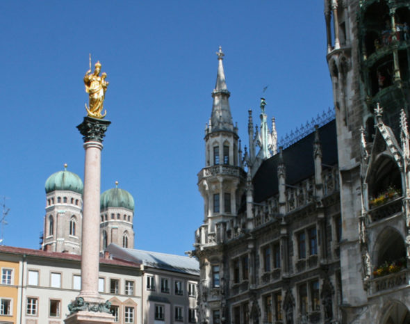 Das Bild ist ein Foto. Es zeigt die Mariensäule auf dem Marienplatz in München. Die Frauenkirche ist im Hintergrund. Der Himmel ist blau.